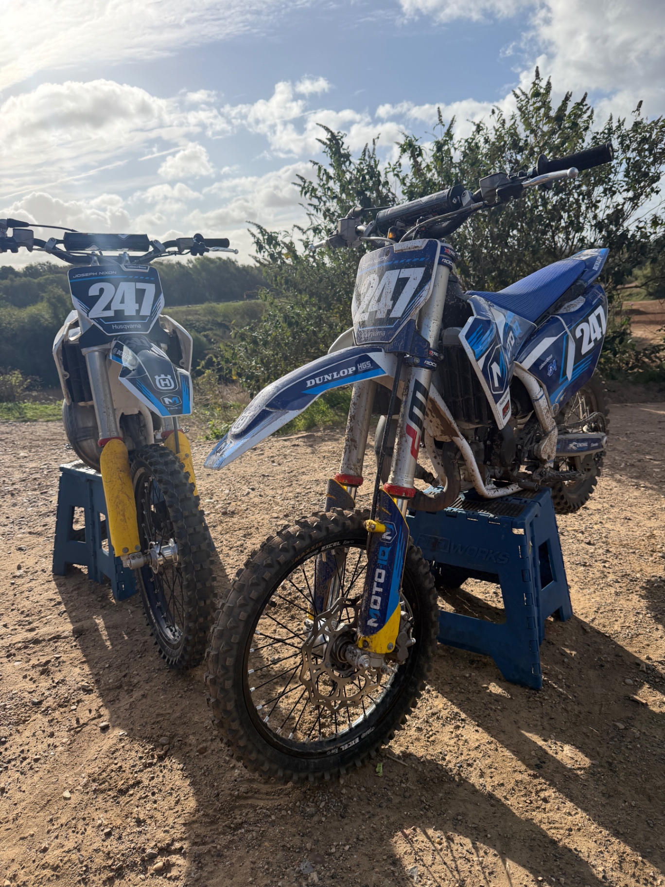 Two blue motocross bikes numbered 247 and 241 standing outdoors beneath a bright sky.