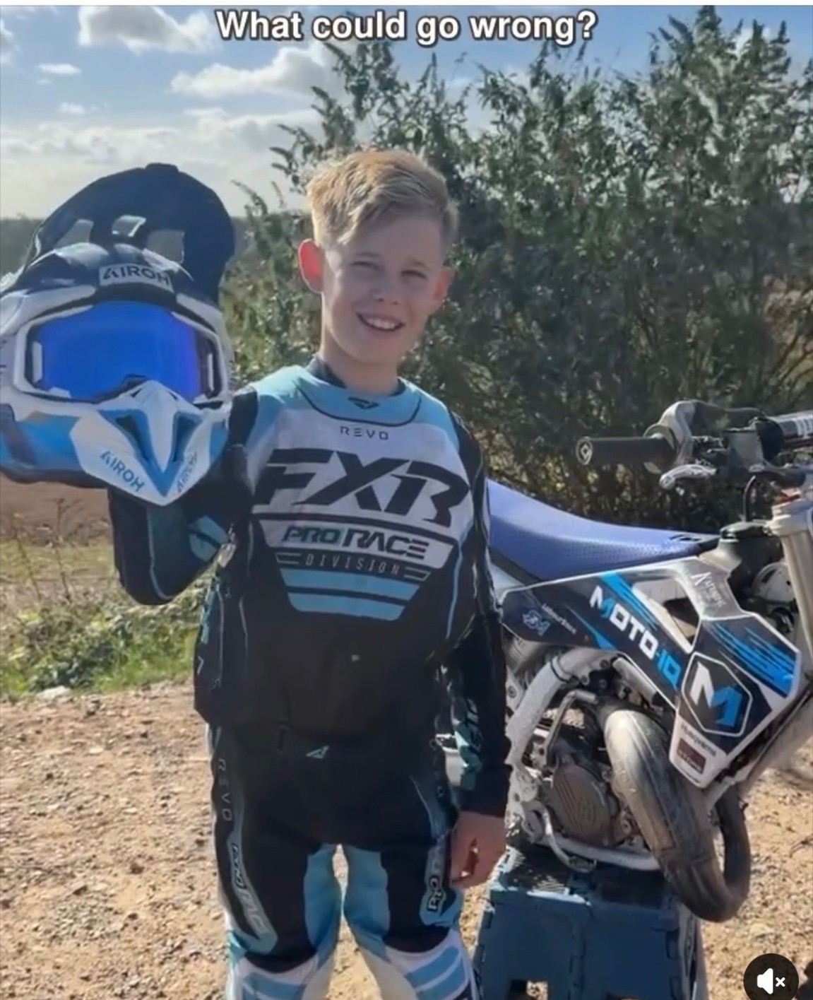 A young motocross rider smiling beside a blue and white dirt bike while holding a helmet.