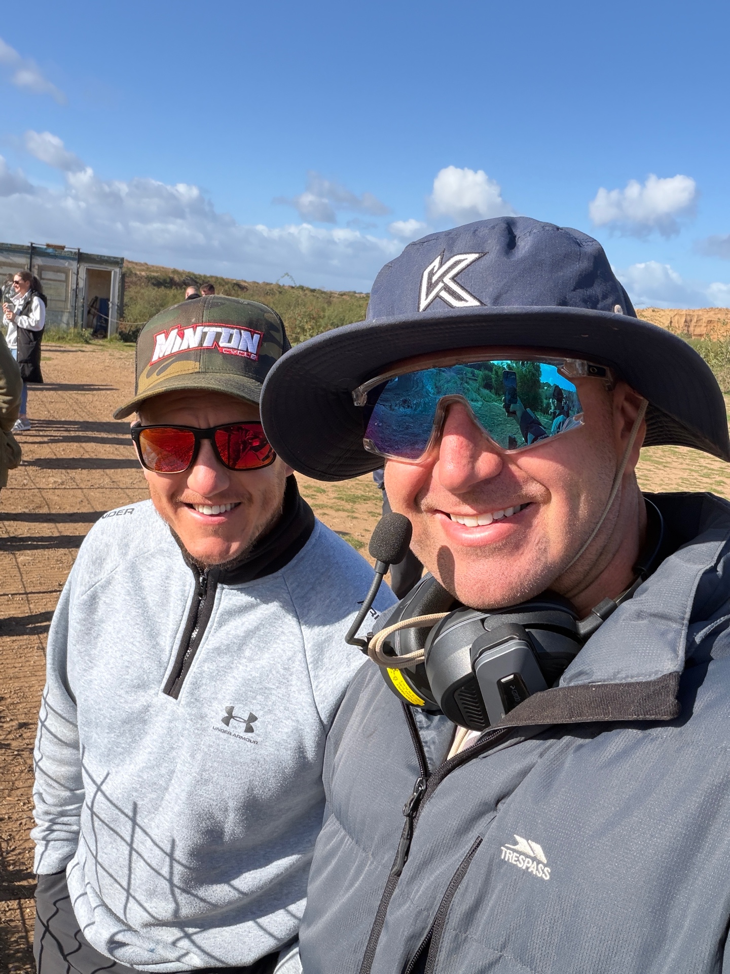 Two smiling motocross crew members wearing caps, sunglasses and communications equipment outdoors.